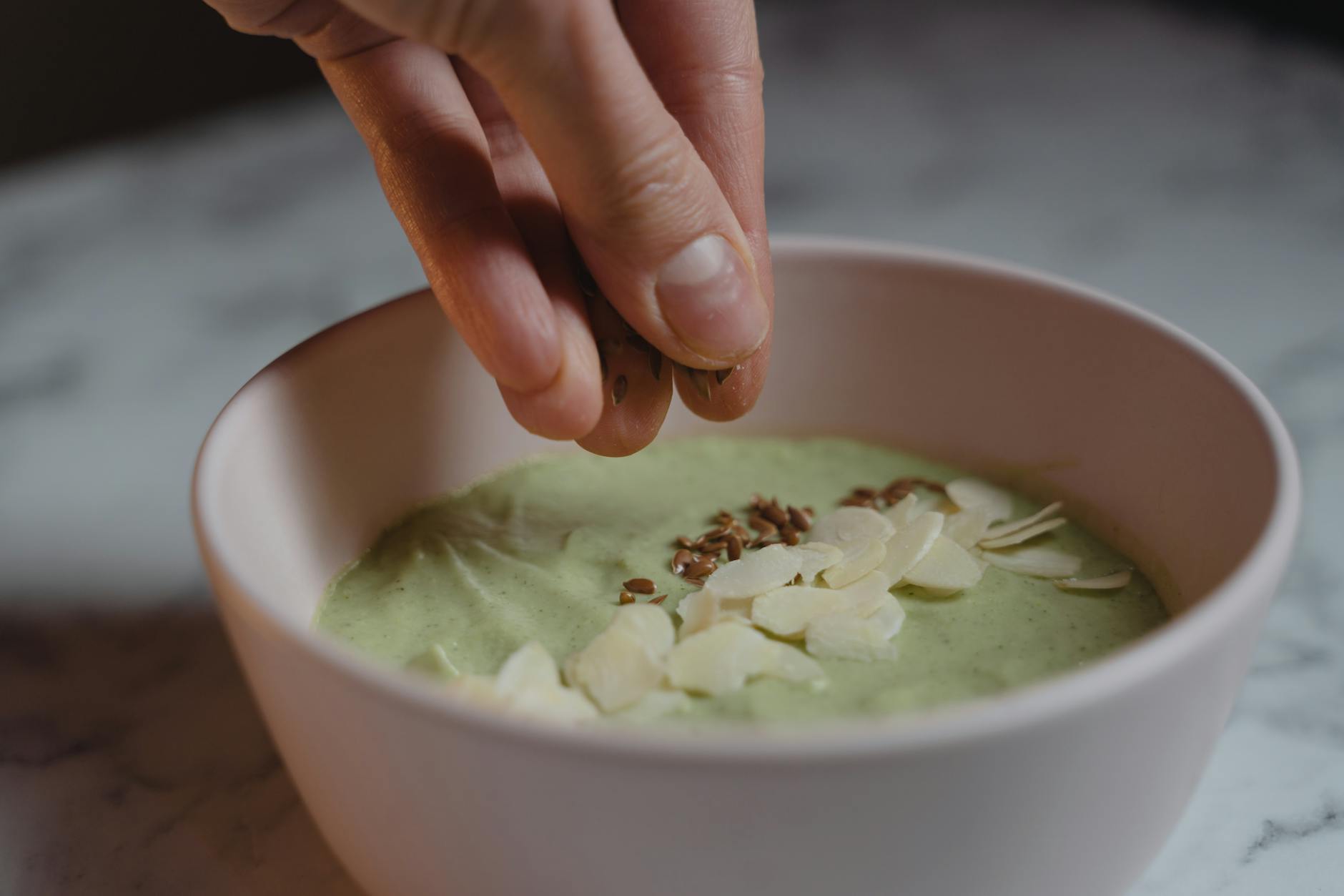 Dłoń dodająca migdały i nasiona do zielonej miski smoothie matcha